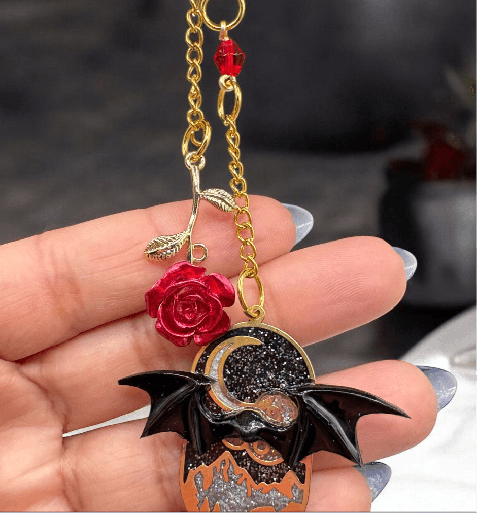 Close-up of ACOTAR bookmark featuring a Velaris charm with bat wings, red rose detail, and gold chain held in hand.